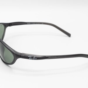 May include: Black Ray-Ban sunglasses with green lenses. The sunglasses have a curved frame and a thin, flexible arm.