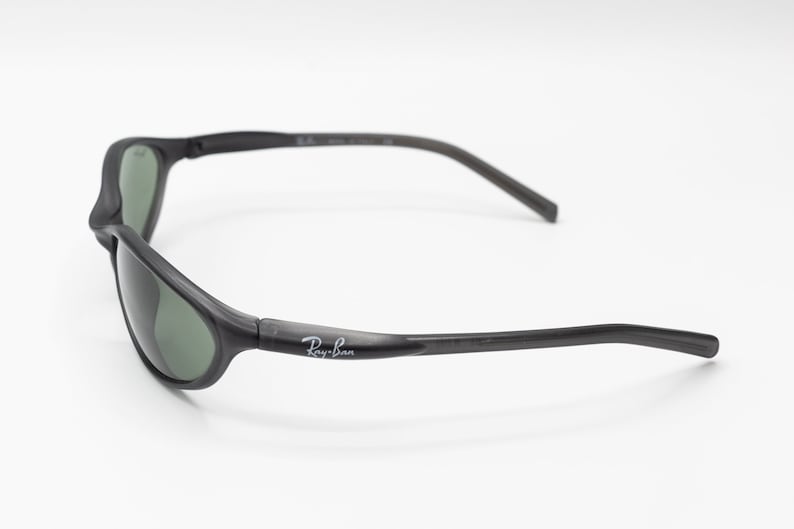 May include: Black Ray-Ban sunglasses with green lenses. The sunglasses have a curved frame and a thin, flexible arm.