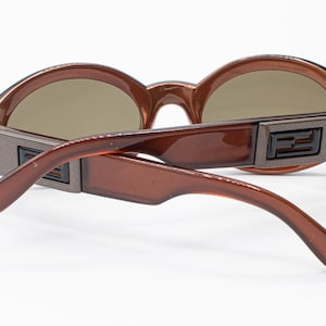 May include: Brown plastic sunglasses with round lenses and a brown and silver metal frame. The sunglasses have a double bridge and the brand name "FENDI" is visible on the frame.