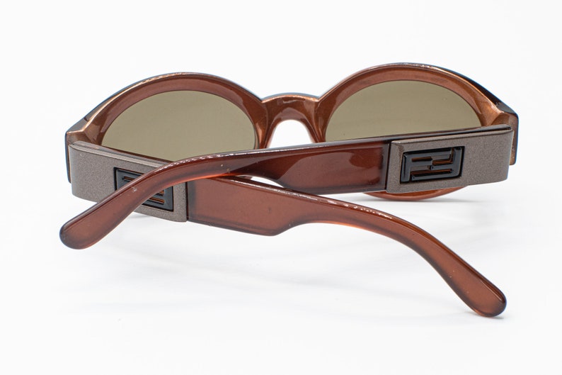 May include: Brown plastic sunglasses with round lenses and a brown and silver metal frame. The sunglasses have a double bridge and the brand name "FENDI" is visible on the frame.