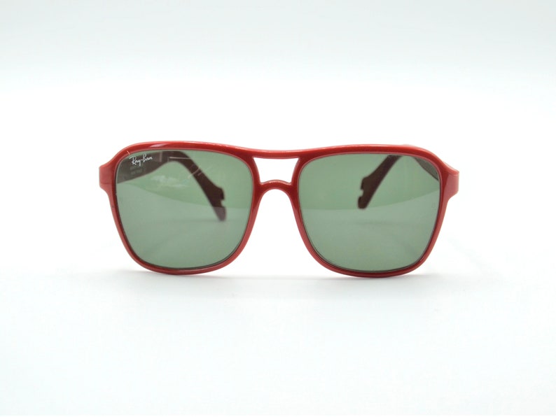 May include: Red plastic aviator sunglasses with green lenses. The sunglasses have the Ray-Ban logo on the left lens.