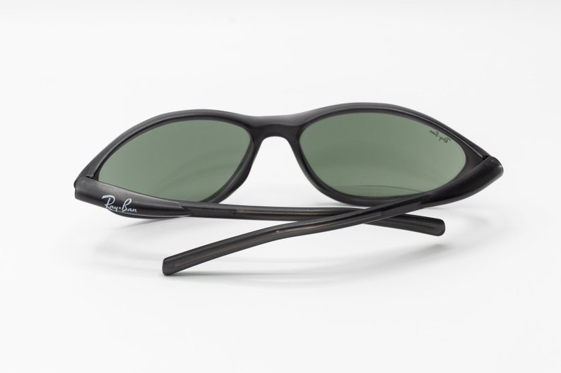 May include: Black Ray-Ban sunglasses with green lenses. The sunglasses have a curved frame and a thin, black temple.