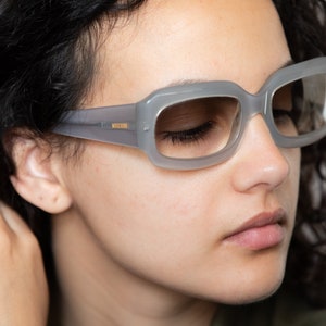 May include: A woman wearing gray rectangular sunglasses with a gold logo on the side. The sunglasses have a light gray frame and clear lenses.