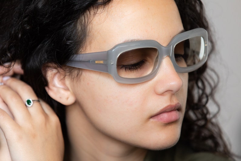 May include: A woman wearing gray rectangular sunglasses with a gold logo on the side. The sunglasses have a light gray frame and clear lenses.