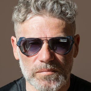 Peut inclure: Lunettes de soleil aviateur avec une monture en métal foncé et des verres dégradés. Les branches sont ornées d'un matériau texturé bleu foncé. Les lunettes ont un petit autocollant rectangulaire sur la lentille avec le texte "100% UV PROTECTION".