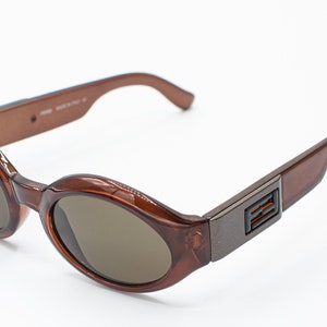 May include: Brown oval-shaped sunglasses with a dark brown lens and a brown plastic frame. The frame has a metal accent with the letters "FENDI" on the side.