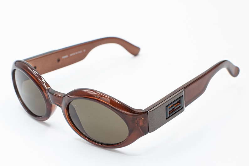 May include: Brown oval-shaped sunglasses with a dark brown lens and a brown plastic frame. The frame has a metal accent with the letters "FENDI" on the side.
