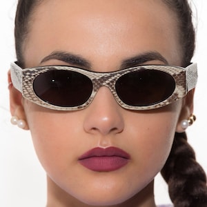 Peut inclure: Une femme portant des lunettes de soleil ovales avec un motif de peau de serpent brun et blanc. Les lunettes ont des verres foncés.