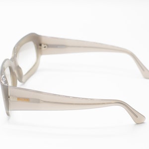 May include: A pair of light gray plastic framed eyeglasses with a rectangular lens shape. The glasses have a subtle curve to the frame and a thin, gold-colored logo on the side.
