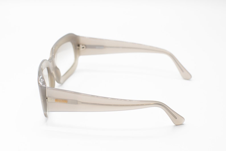 May include: A pair of light gray plastic framed eyeglasses with a rectangular lens shape. The glasses have a subtle curve to the frame and a thin, gold-colored logo on the side.