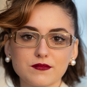 Puede incluir: Una mujer con gafas de montura dorada con el nombre de la marca "Salvatore Ferragamo" impreso en la lente derecha. Tiene el pelo castaño oscuro, los ojos castaño oscuro y lleva lápiz labial rojo.