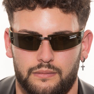 Peut inclure: Un homme portant une veste en cuir noire et des lunettes de soleil avec une monture à imprimé camouflage. Les lunettes de soleil ont des verres foncés et le texte "100% UV PROTECTION" et "Roberto Cavalli" sont imprimés sur la lentille droite.