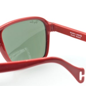 May include: Red aviator sunglasses with green lenses. The sunglasses have a metal frame and the text "BAUSCH & LOMB FRAME FRANCE" is printed on the side.