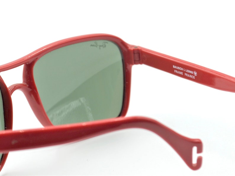 May include: Red aviator sunglasses with green lenses. The sunglasses have a metal frame and the text "BAUSCH & LOMB FRAME FRANCE" is printed on the side.