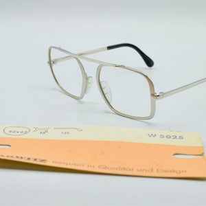 Puede incluir: Gafas de metal plateadas con montura rectangular y patillas de pl&aacute;stico negro. Las gafas est&aacute;n sobre una superficie blanca con una peque&ntilde;a etiqueta que dice "W 5025" y "exquisite in Qualitat und Design".