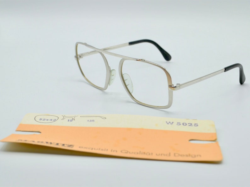 Puede incluir: Gafas de metal plateadas con montura rectangular y patillas de pl&aacute;stico negro. Las gafas est&aacute;n sobre una superficie blanca con una peque&ntilde;a etiqueta que dice "W 5025" y "exquisite in Qualitat und Design".