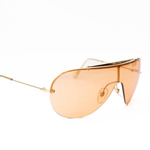 K&ouml;nnte beinhalten: Sonnenbrille mit goldfarbenem Rahmen und gro&szlig;em Einzellinsen-Design in Pfirsicht&ouml;nung. Die Sonnenbrille hat eine schlanke, moderne &Auml;sthetik mit einem d&uuml;nnen Goldrahmen und einem geschwungenen B&uuml;gel. Die Gl&auml;ser sind einfarbig.