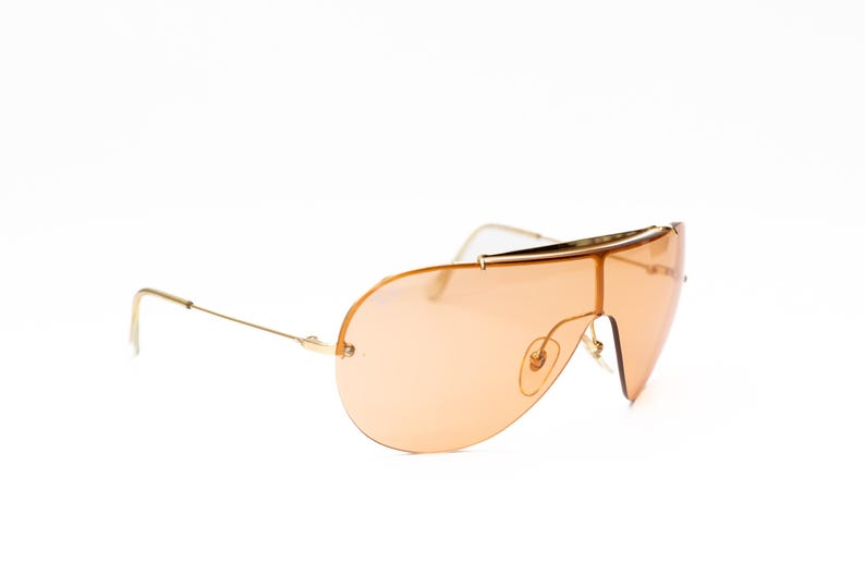 K&ouml;nnte beinhalten: Sonnenbrille mit goldfarbenem Rahmen und gro&szlig;em Einzellinsen-Design in Pfirsicht&ouml;nung. Die Sonnenbrille hat eine schlanke, moderne &Auml;sthetik mit einem d&uuml;nnen Goldrahmen und einem geschwungenen B&uuml;gel. Die Gl&auml;ser sind einfarbig.