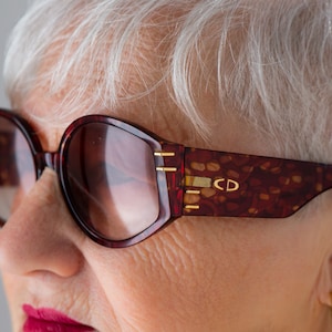 Puede incluir: Primer plano de una mujer con gafas de sol de carey rojo con detalles dorados y las letras "CD" en el lateral.