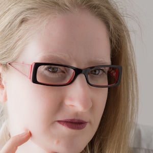 May include: A woman with blonde hair wearing black framed glasses with red accents. She is wearing a white shirt and has a serious expression.