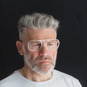 Puede incluir: Un hombre con cabello gris y barba lleva gafas de montura blanca. Est&aacute; mirando hacia abajo y tiene una expresi&oacute;n seria en su rostro.