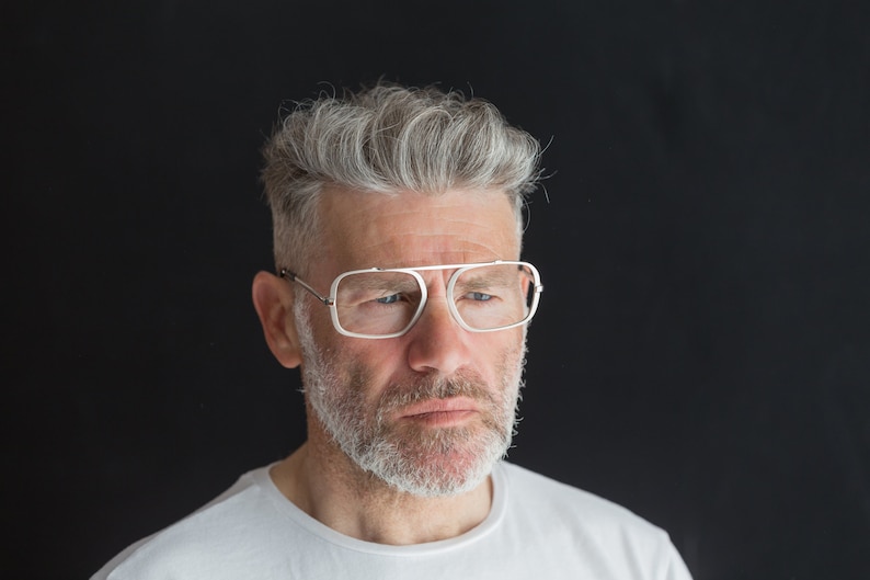 Puede incluir: Un hombre con cabello gris y barba lleva gafas de montura blanca. Est&aacute; mirando hacia abajo y tiene una expresi&oacute;n seria en su rostro.