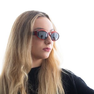 May include: A woman with long blonde hair wearing oval-shaped sunglasses with a red frame and dark lenses.