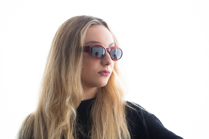 May include: A woman with long blonde hair wearing oval-shaped sunglasses with a red frame and dark lenses.