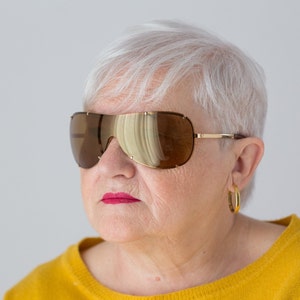 May include: A woman with short, white hair wearing large, brown, wraparound sunglasses with gold frames. She is wearing a yellow sweater.