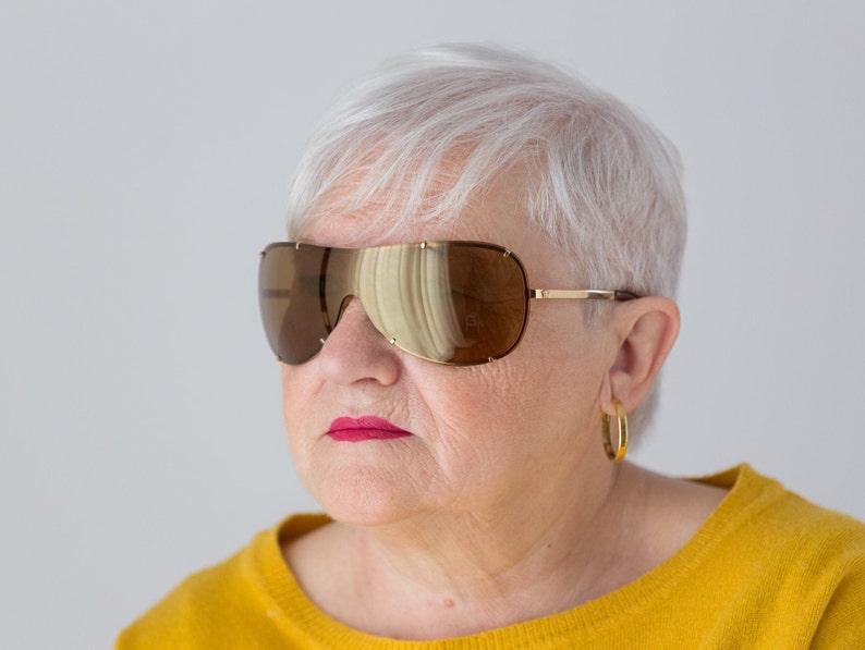 May include: A woman with short, white hair wearing large, brown, wraparound sunglasses with gold frames. She is wearing a yellow sweater.