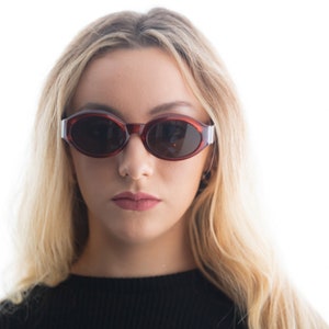 May include: A woman with long blonde hair wearing oval-shaped sunglasses with a red frame and dark lenses.