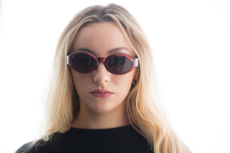 May include: A woman with long blonde hair wearing oval-shaped sunglasses with a red frame and dark lenses.