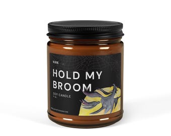 May include: A soy candle in a glass jar with a black lid. The label reads "HOLD MY BROOM" with a bat illustration. The candle is 9 oz.