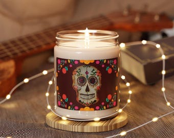 May include: A clear glass jar candle with a white wax fill and a lit wick. The label features a central sugar skull illustration surrounded by colorful floral patterns on a dark brown background, resting on a circular wooden base.