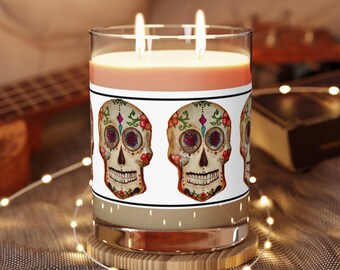 May include: A clear glass candle with two lit wicks and a light orange wax fill. The exterior features a white label decorated with a repeating pattern of colorful sugar skull illustrations, each adorned with floral and geometric details.