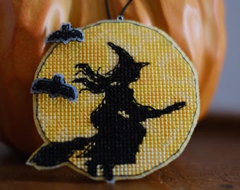 May include: A circular cross-stitch ornament featuring a black silhouette of a witch flying on a broomstick against a yellow moon background. Two small, dark grey bat-shaped embellishments are attached to the left side. A thin black string is attached.