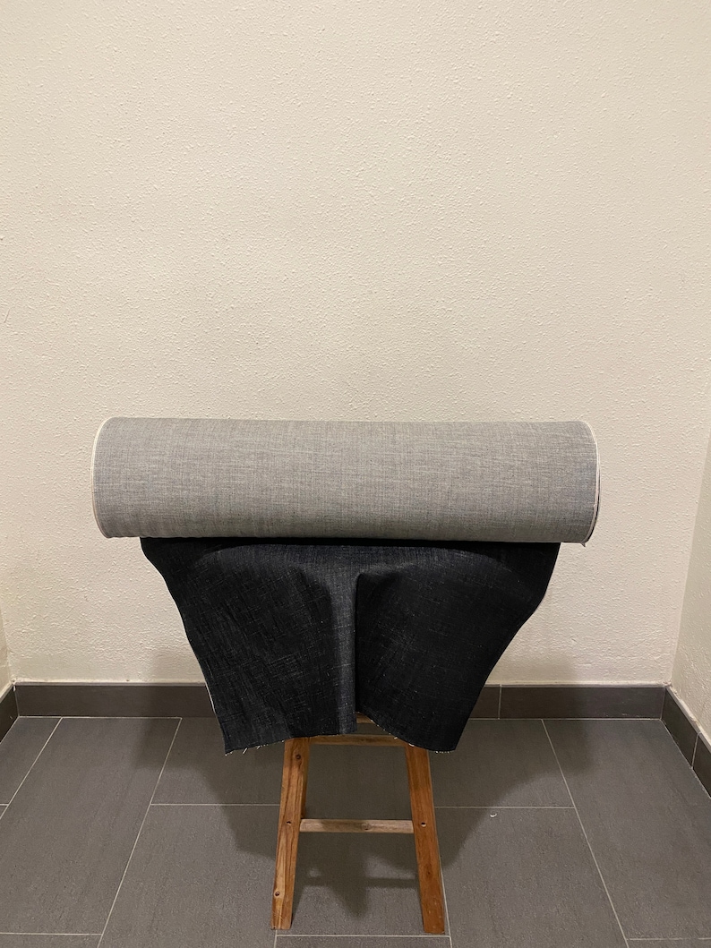 May include: A roll of gray fabric and a roll of black fabric are displayed on a wooden stool. The fabric is folded and placed on top of the stool.