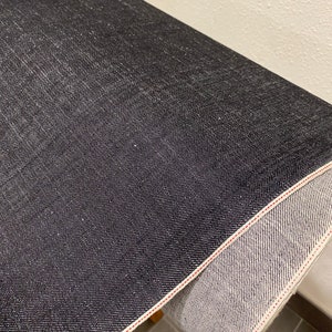 May include: Close-up of a dark blue denim fabric with a red stitching detail. The fabric is rolled up and the inside of the roll is visible.