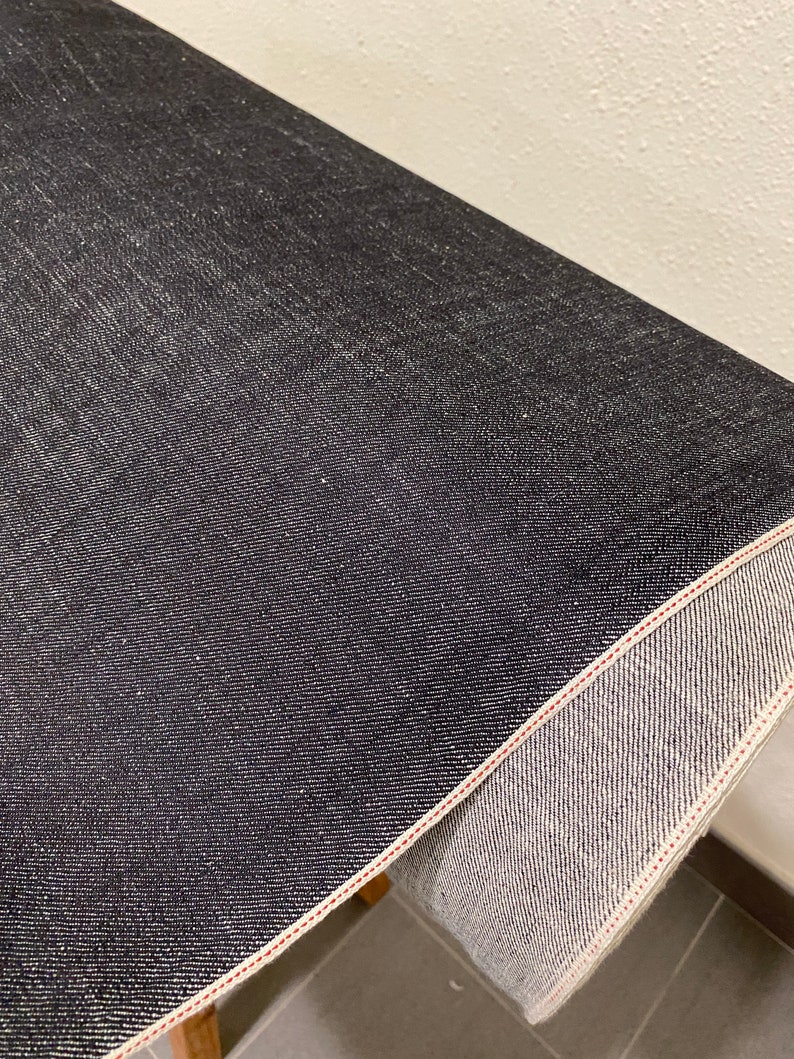 May include: Close-up of a dark blue denim fabric with a red stitching detail. The fabric is rolled up and the inside of the roll is visible.