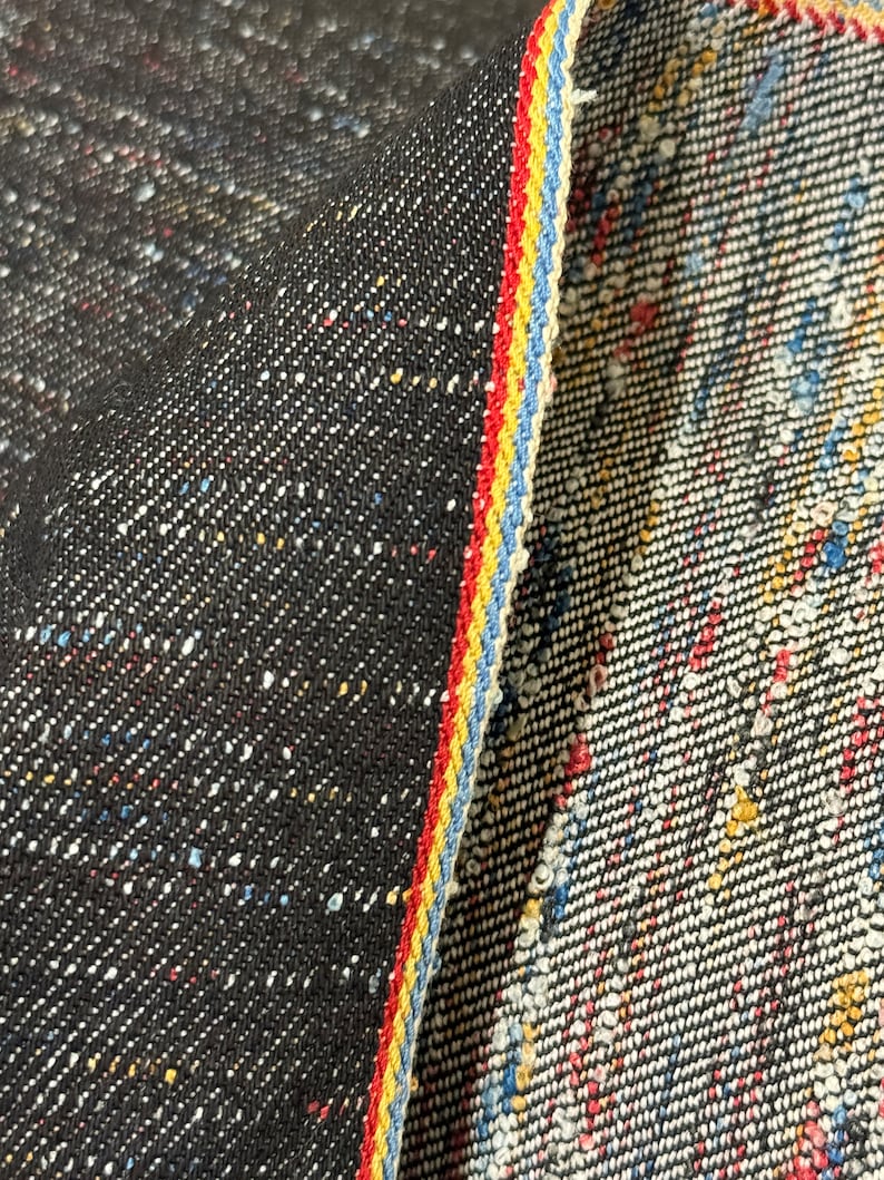 May include: Black denim fabric with a colorful speckled pattern. The fabric has a red, yellow, and blue selvedge edge.