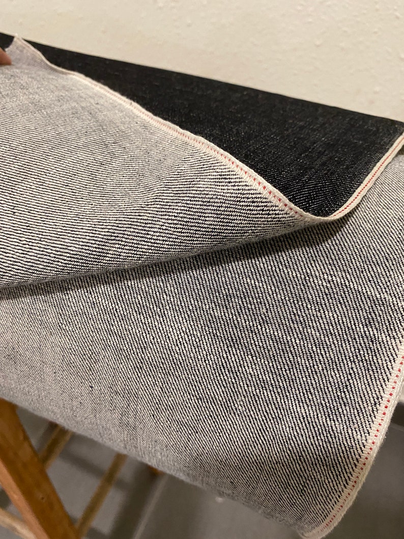 May include: Close-up of black denim fabric with a red selvedge edge. The fabric is folded to reveal the white weft threads.
