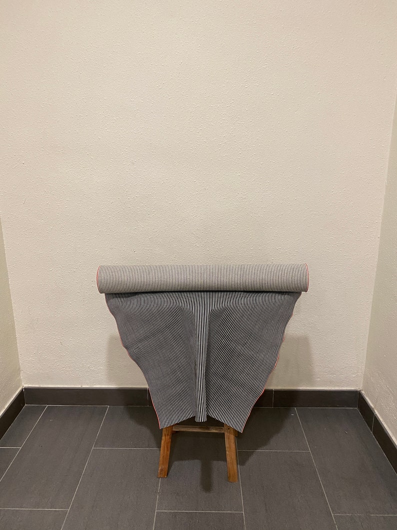 May include: A roll of black and white striped fabric with a red edge is displayed on a wooden stool. The fabric is rolled up and the edges are visible.