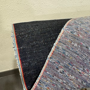 May include: Close-up of two different types of denim fabric. One is a dark blue denim with a subtle texture. The other is a light blue denim with a multi-colored woven pattern. Both fabrics have a red, white, and blue selvedge edge.