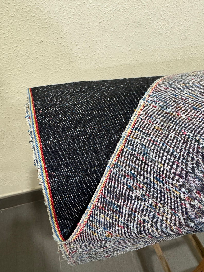 May include: Close-up of two different types of denim fabric. One is a dark blue denim with a subtle texture. The other is a light blue denim with a multi-colored woven pattern. Both fabrics have a red, white, and blue selvedge edge.