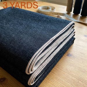 Może przedstawiać: Zestaw dżinsowy z denimu z napisem "JEANS KIT 3 YARDS". Ciemnoniebieski materiał dżinsowy jest złożony, odsłaniając jasną krawędź. W tle widoczne są akcesoria do szycia, w tym szpulki nici i miarka.