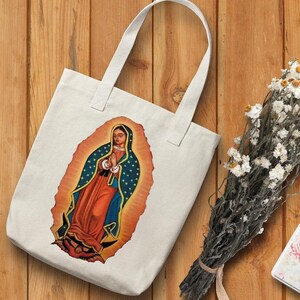 May include: A white canvas tote bag with a colorful image of the Virgin Mary of Guadalupe. The image is surrounded by a golden halo and stars.