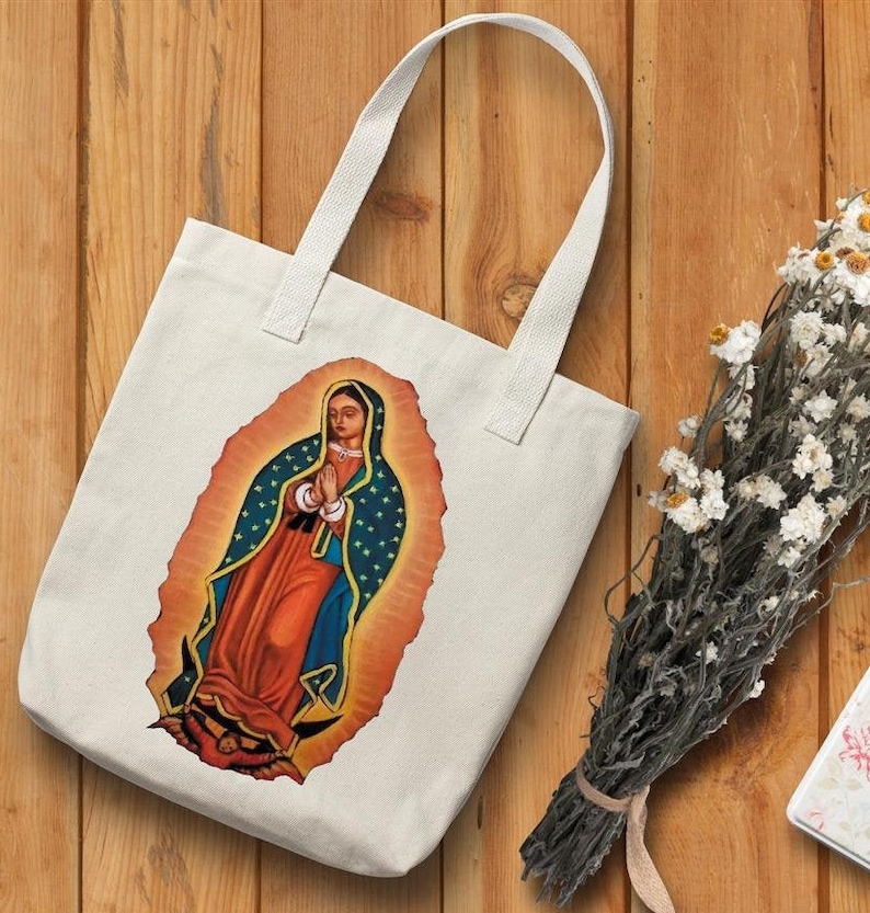 May include: A white canvas tote bag with a colorful image of the Virgin Mary of Guadalupe. The image is surrounded by a golden halo and stars.