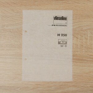May include: A rectangular sheet of Vlieseline H 250 interfacing, with black printed branding and product details. The sheet is a light beige colour and has a textured surface. It is placed on a light brown wooden surface.