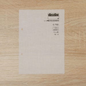 Puede incluir: Una muestra de tela rectangular de color blanquecino con un borde perforado y dos agujeros. La muestra presenta el texto "Vlieseline" y "G 700", junto con símbolos de cuidado para planchar y lavar. La tela está sobre una superficie de madera clara.