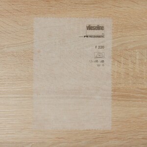 Puede incluir: Una lámina rectangular translúcida de entretela Vlieseline Freudenberg F 220. La lámina presenta texto y símbolos impresos, incluido el nombre de la marca, el código del producto y las instrucciones de cuidado. Fondo de veta de madera clara.